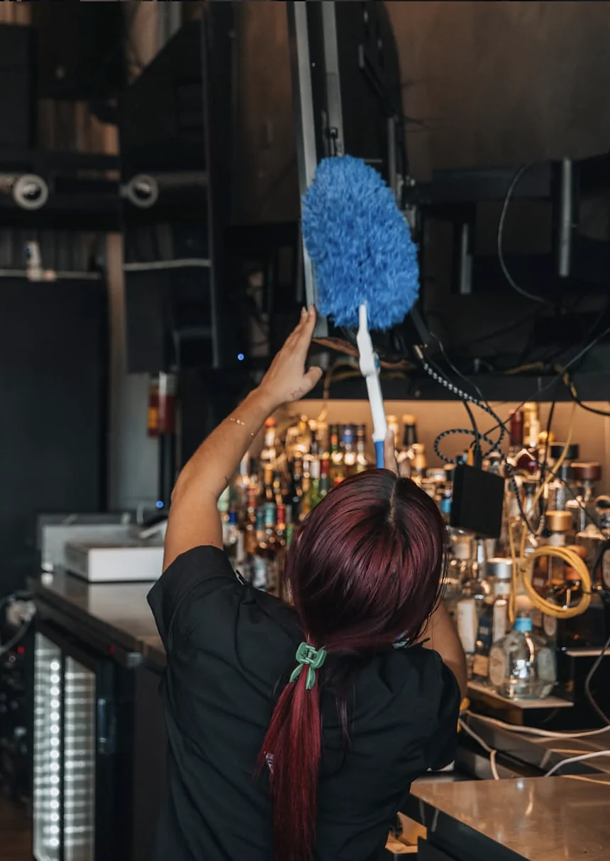 Empleada de Clean Vibes desempolvando la estantería de botellas de una barra