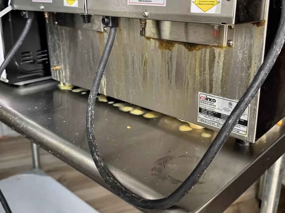 Freidora industrial con grasa acumulada escurriendo por el frente y residuos sobre la mesa de acero