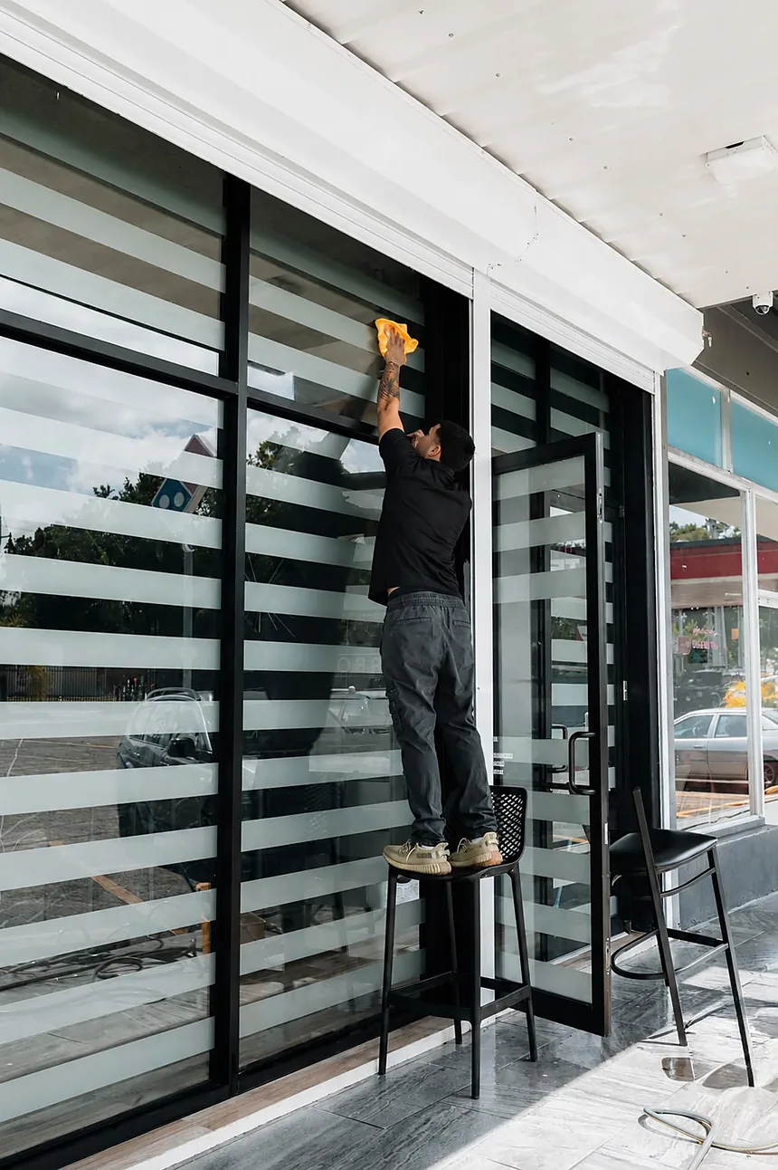 Empleado de Clean Vibes subido en un banquito limpiando los cristales altos de la fachada de un local comercial